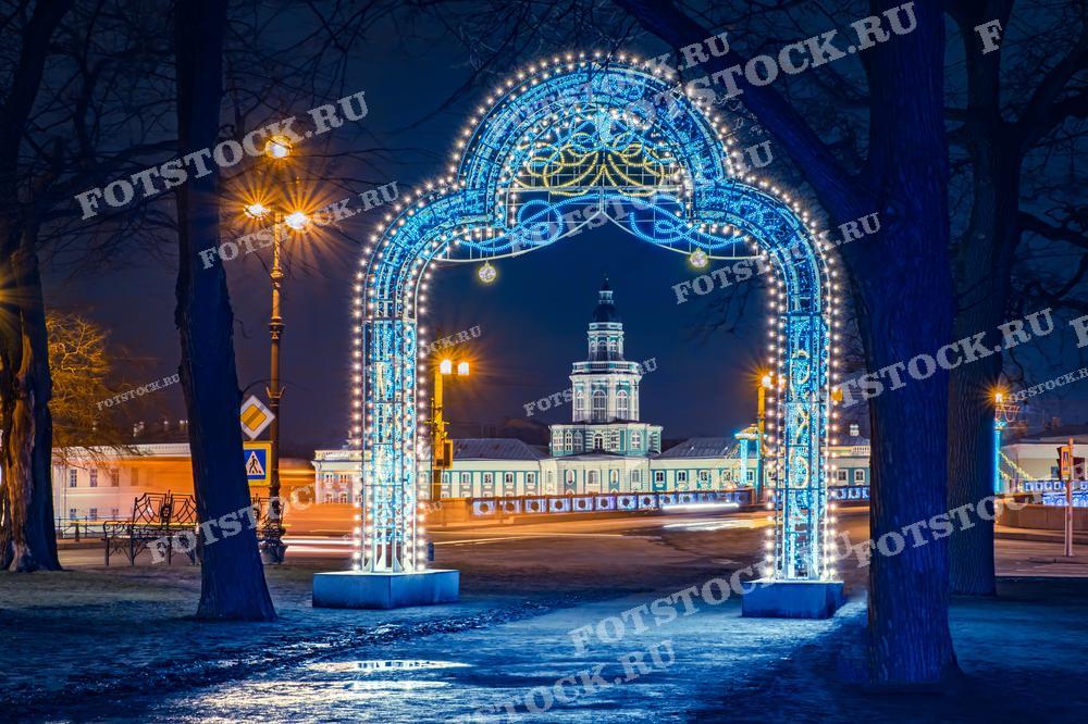 На фотографии изображена новогодняя арка, украшенная гирляндами, с видом на Кунсткамеру ночью в Санкт-Петербурге. Снимок отлично подходит для использования в материалах, посвященных рождественским и новогодним праздникам, а также для туристических буклетов и сайтов.